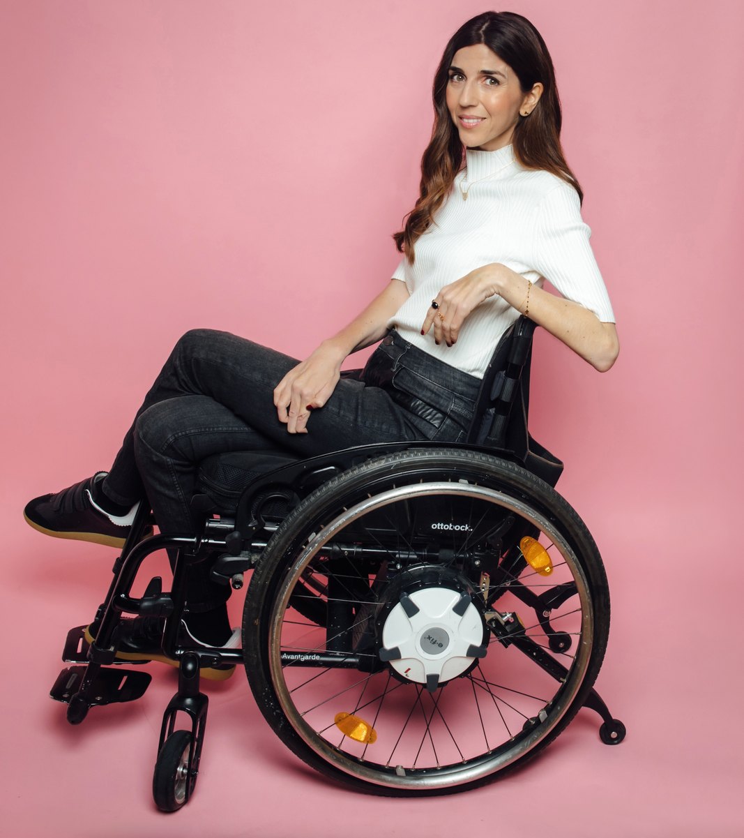 Virginie Dubost, souriante, dans son fauteuil roulant, sur fond rose.