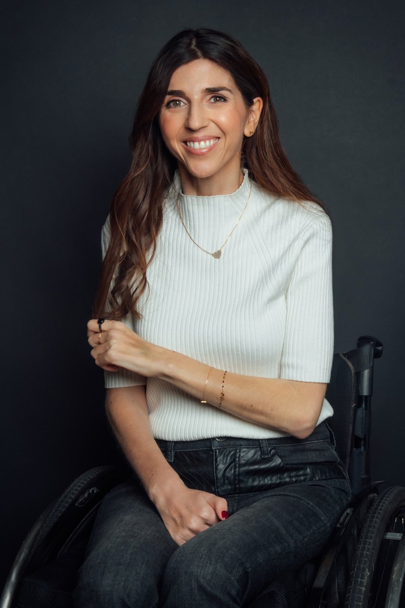 Portrait de Virginie Dubost, souriante, dans son fauteuil roulant, sur fond sombre.